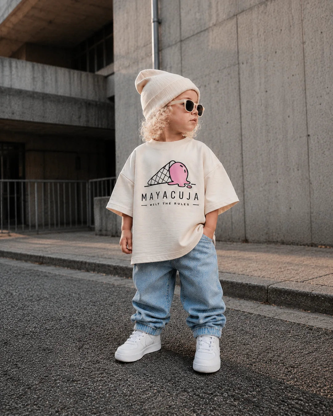toddler in an oversized cream mayacuja tee and light denim, standing on asphalt in front of a concrete wall